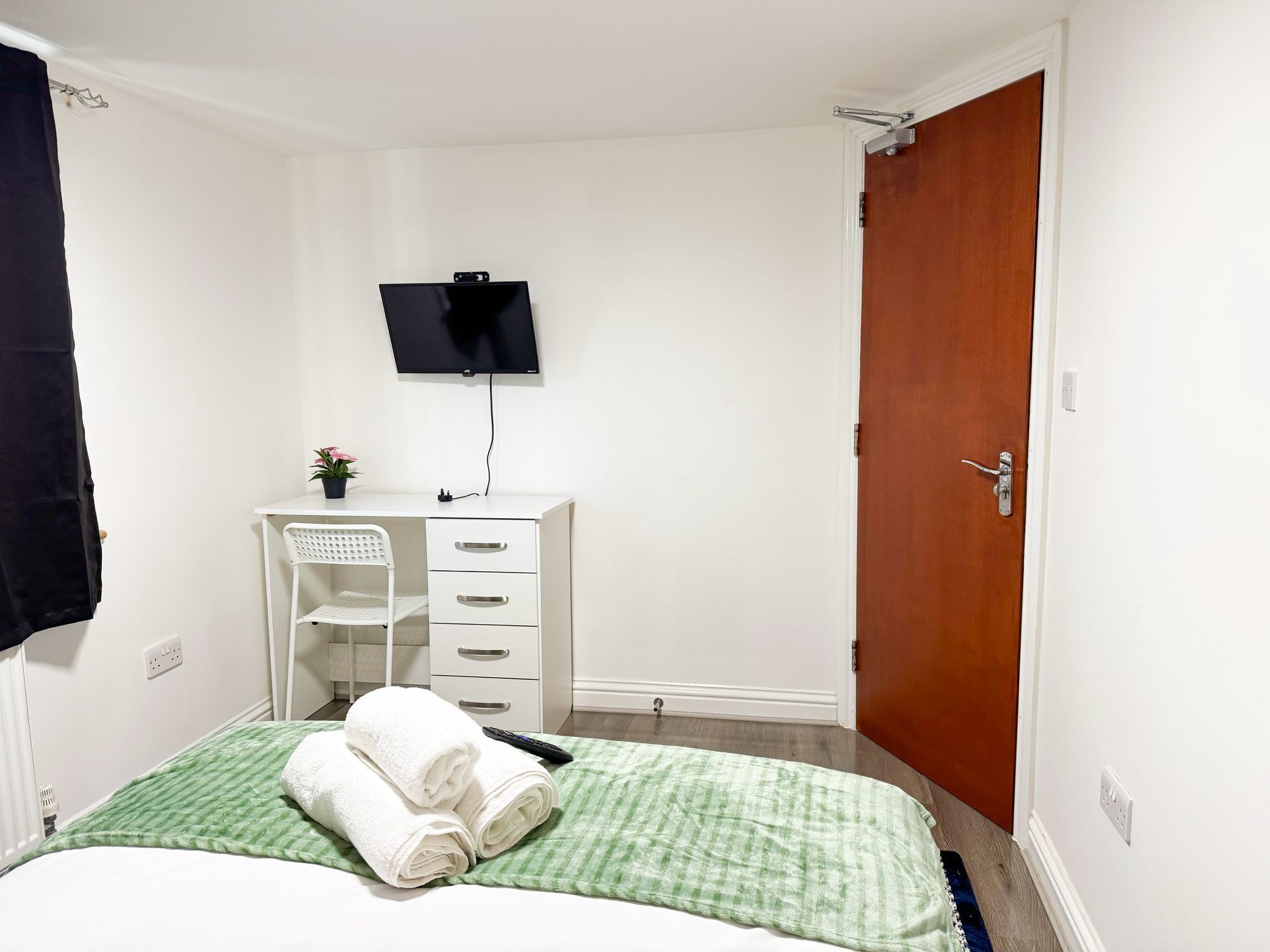 A hotel room with a bed, a desk with a chair, a television mounted on the wall, and a wooden door.