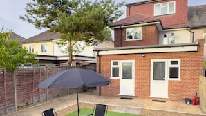 A patio with a round table and four chairs, an umbrella, and a brick house in the background. - Heathrow Lodge B&B (Hayes)