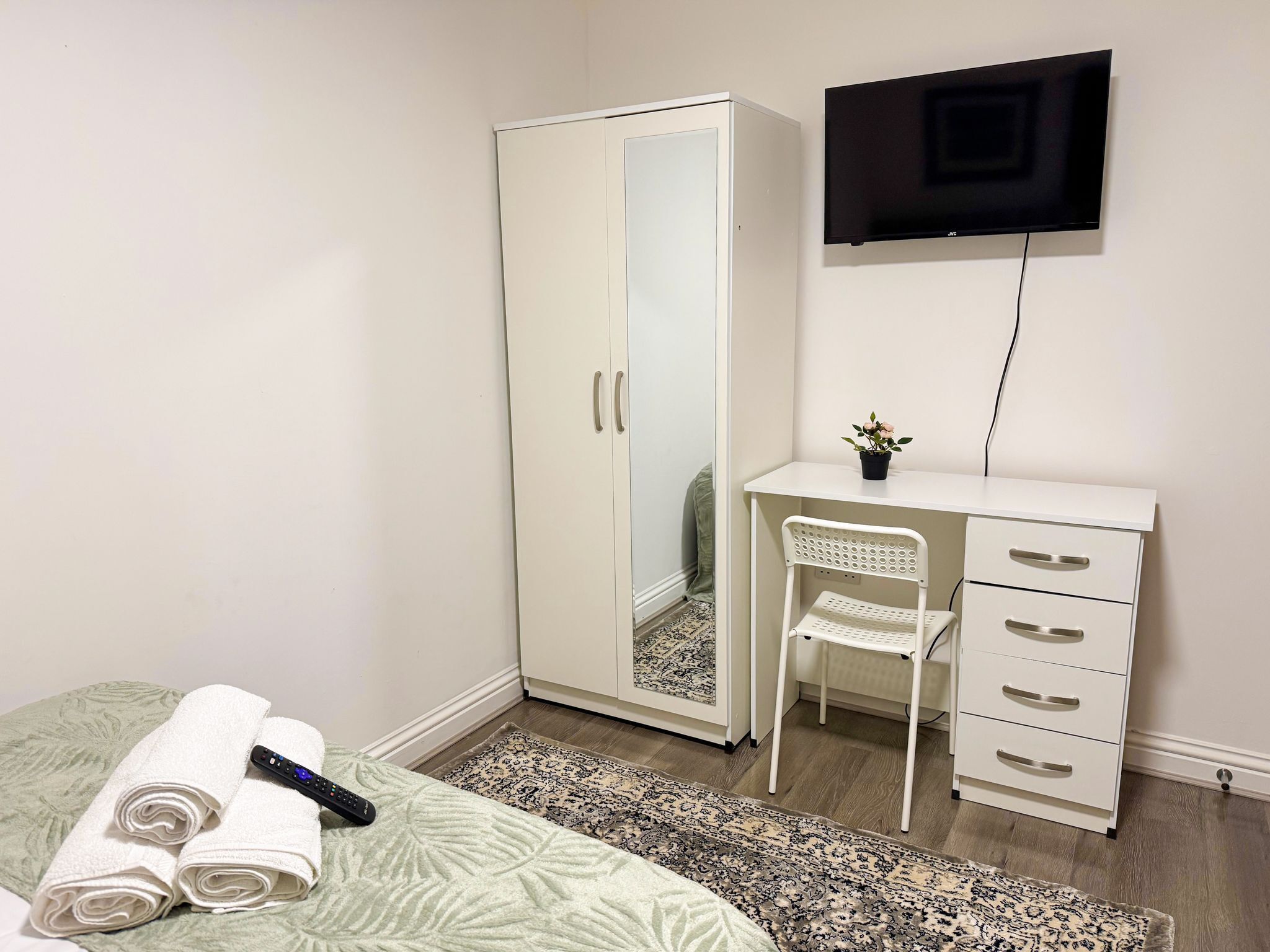 A bedroom with a bed, a wardrobe, a desk with a chair, a TV mounted on the wall, and a small potted plant on the desk.