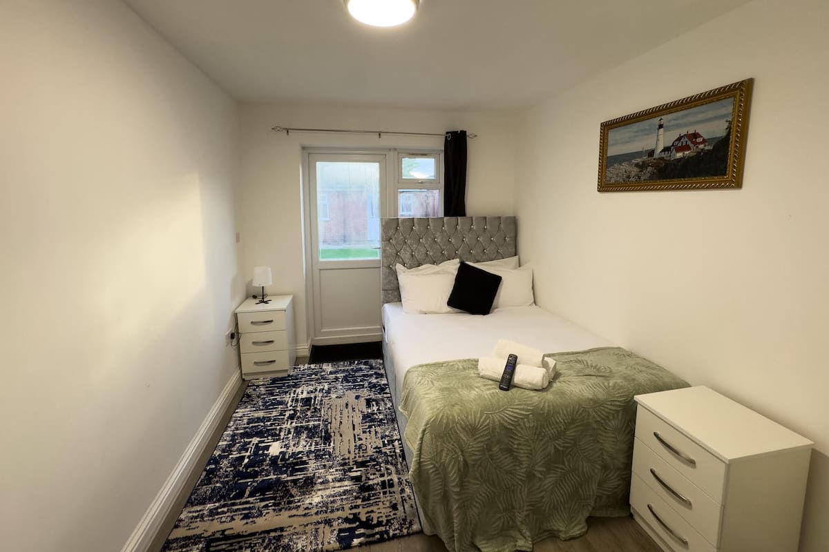 A narrow hotel room with a bed, a bedside table, a small drawer unit, a patterned rug, and a painting on the wall.