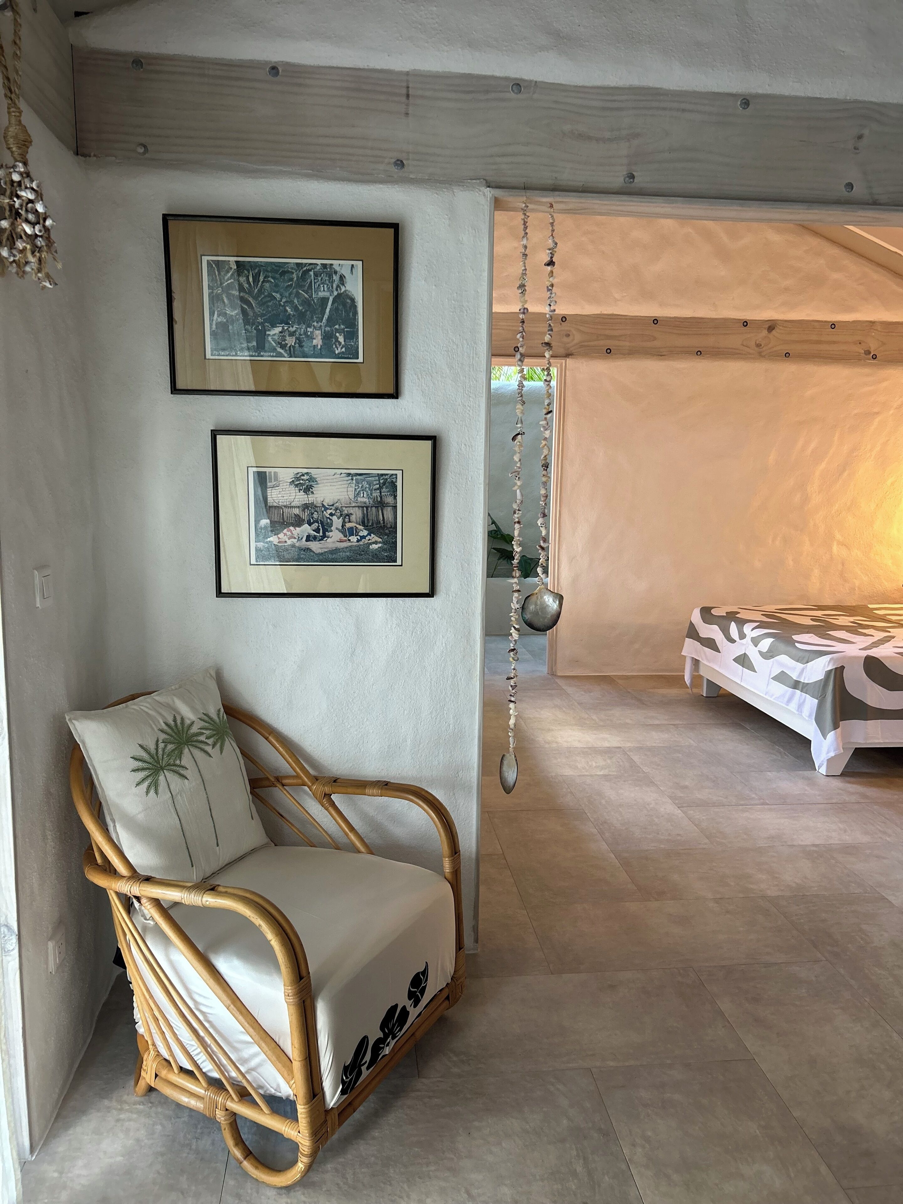 Une pièce comprenant une chaise en osier, deux photographies encadrées accrochées au mur, un objet décoratif suspendu et un lit recouvert d’une couvre-lit à motifs.