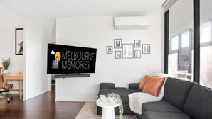 Uma sala de estar moderna com um sofá cinzento, uma mesa de centro branca e uma TV de tela plana fixada na parede exibindo 'Melbourne Memories'.
