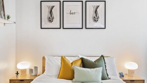 Cama bem arrumada com manta amarela, travesseiros brancos e diversos travesseiros coloridos. Há três quadros emoldurados na parede acima da cama, e duas luminárias de cabeceira de cada lado.