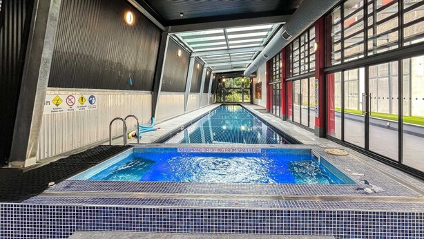 Piscina interna com aviso claro proibindo saltos ou mergulhos, piso ladrilhado e teto de vidro que permite entrada de luz natural.