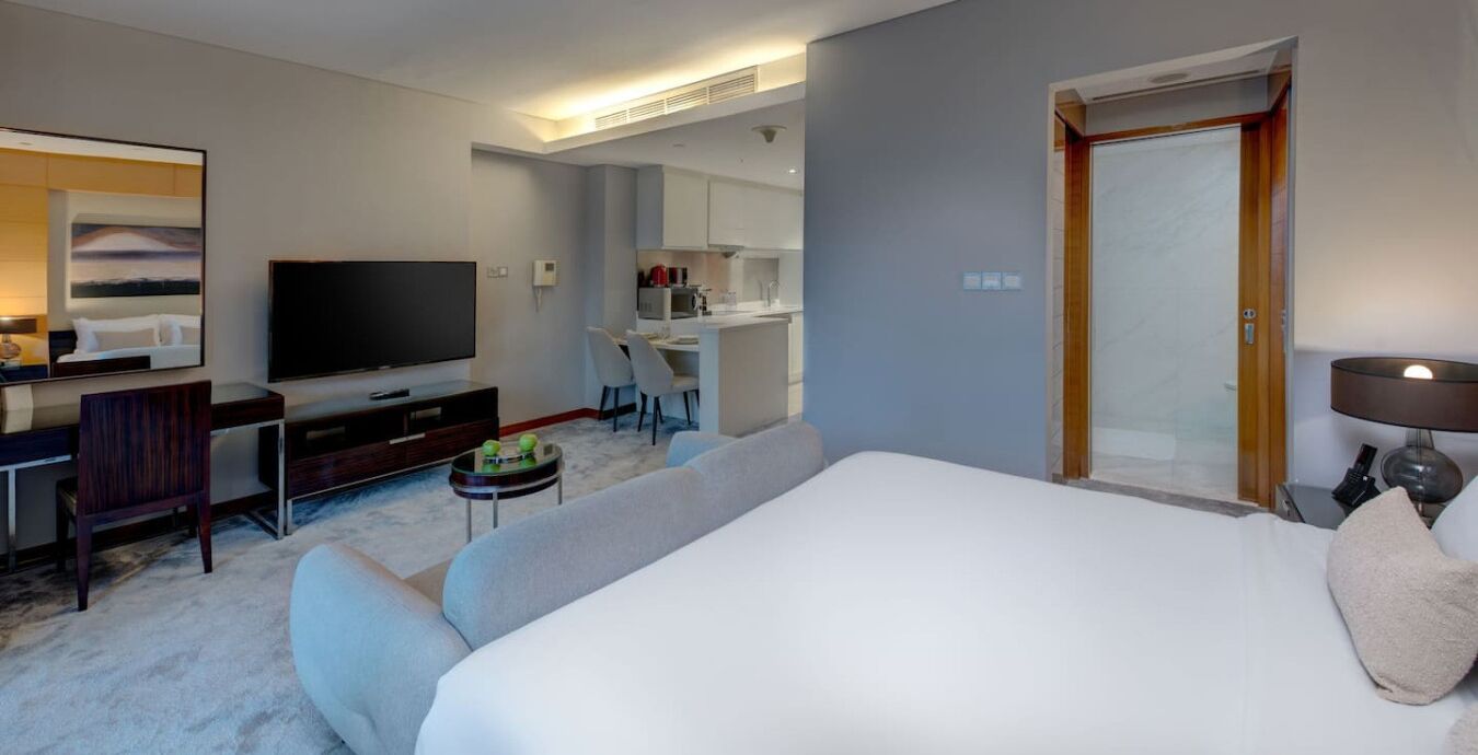 A hotel room with a large bed, a flat-screen TV, a desk with a chair, a small round table with a lamp, and a painting on the wall.