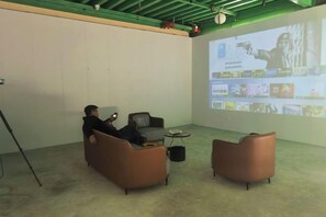 A person using a laptop in a room with a projector screen displaying a presentation, two sofas, and a coffee table.