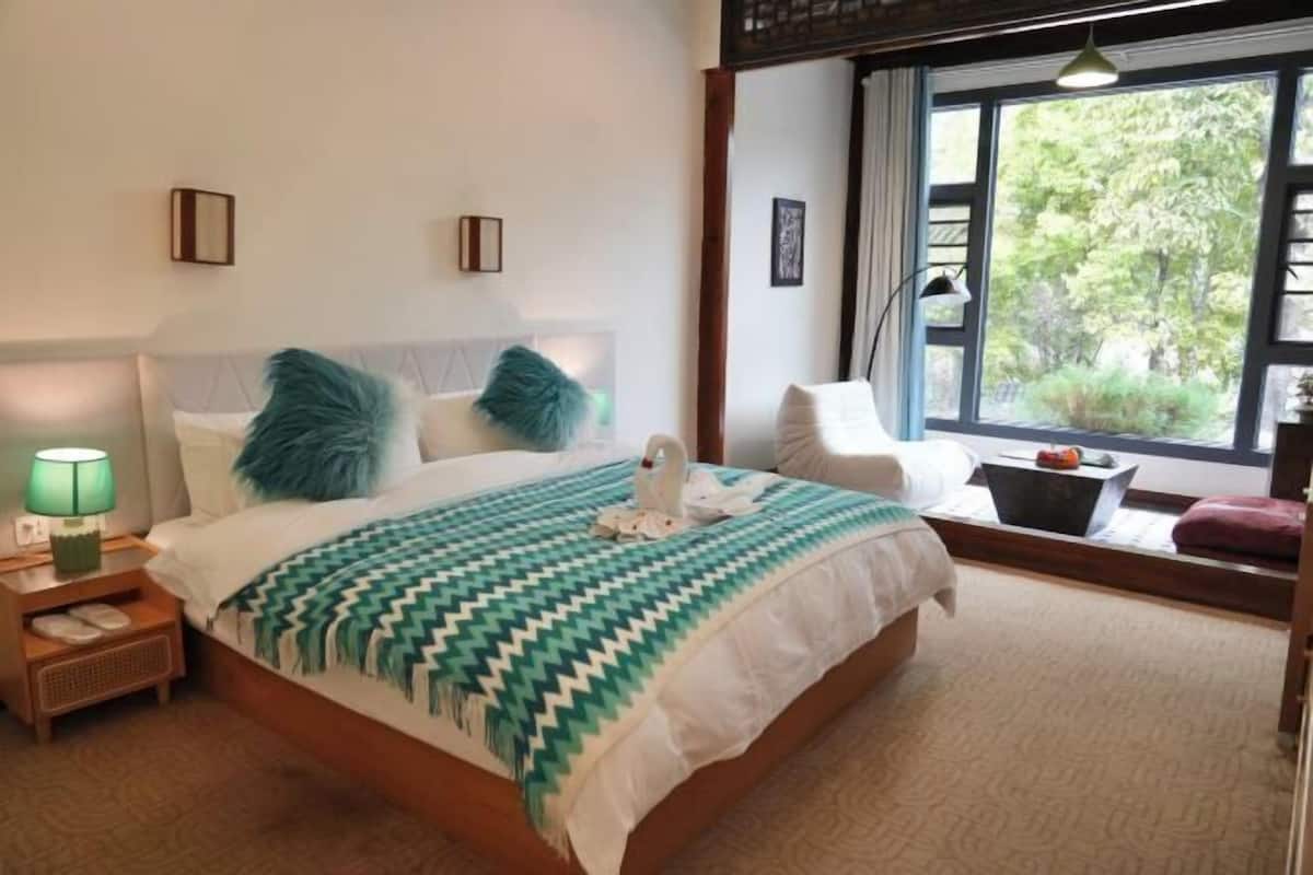 Un dormitorio con cama, mesita de noche, lámpara, ventana con vista a árboles y una silla.