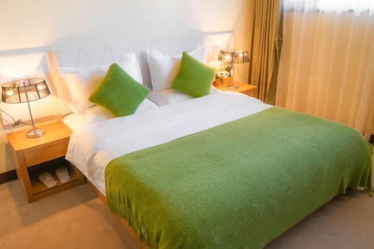 Una habitación de hotel con cama, almohadas, mesita de noche con lámpara y ventana con cortinas.