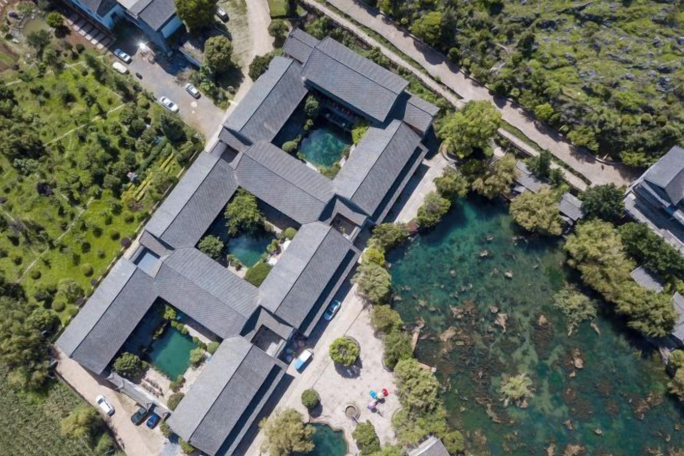 An aerial view of a building with multiple rectangular structures, surrounded by greenery and a body of water.