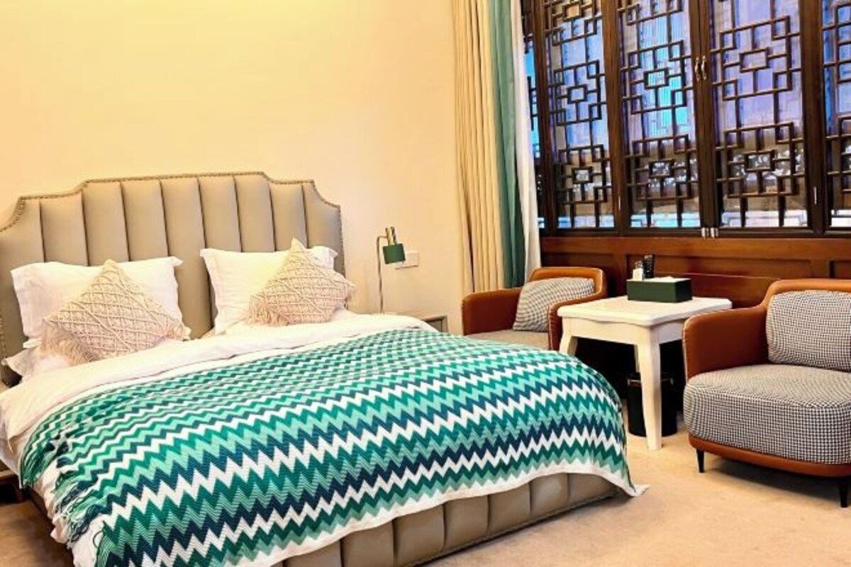 A hotel room with a bed, a small table, two chairs, and a window with a geometric pattern.