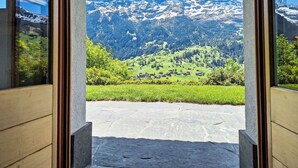 Une vue à travers l’encadrement d’une porte, donnant sur un paysage montagneux avec des sommets enneigés, des vallées verdoyantes et des maisons dispersées.