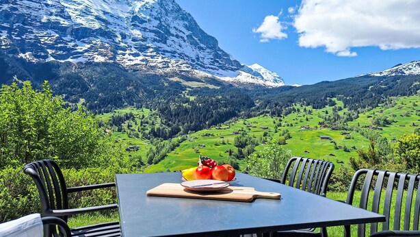 Une table avec une planche à découper, une assiette et deux pommes, le tout placé sur fond d’un paysage montagneux sous un ciel dégagé.