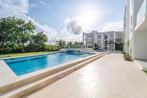 Piscina com degraus de acesso, cercada por um espaço pavimentado e um edifício com várias varandas.