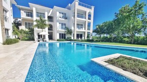 Un hotel moderno con una piscina rectangular, rodeado de exuberante vegetación y un cielo azul despejado.
