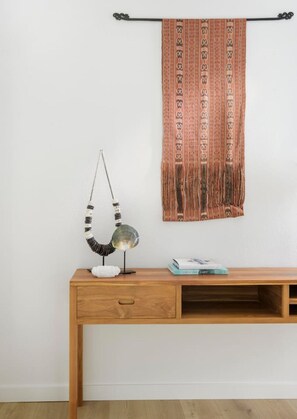 Ein Holztisch mit Lampe und Buch, an einer schlichten Wand mit einem gemusterten Wandteppich.