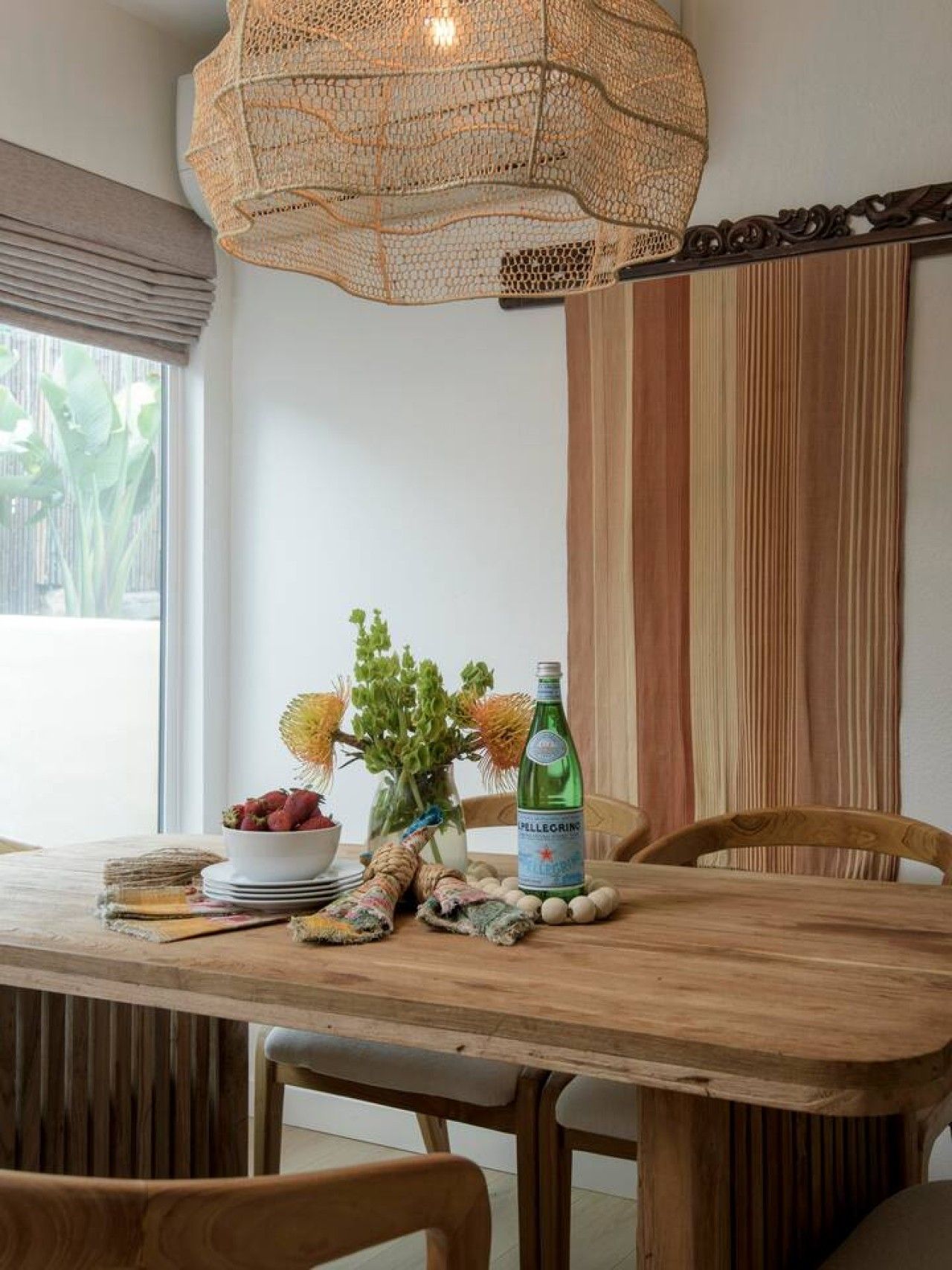 Een houten eettafel met een geweven hanglamp erboven, een fles Pellegrino water en een vaas met groene planten.