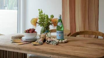 Een houten eettafel met een geweven hanglamp erboven, een fles Pellegrino water en een vaas met groene planten.