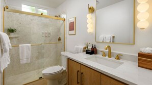 Een badkamer met een douchecabine van glas, een wit toilet, een grote spiegel met wandlampen en een houten wastafelmeubel met een wastafel.