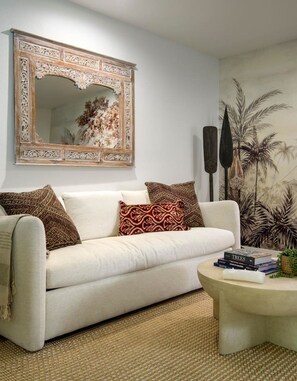 A living room with a white sofa, patterned cushions, a coffee table with books, a large mirror with an ornate frame, and a wall mural.