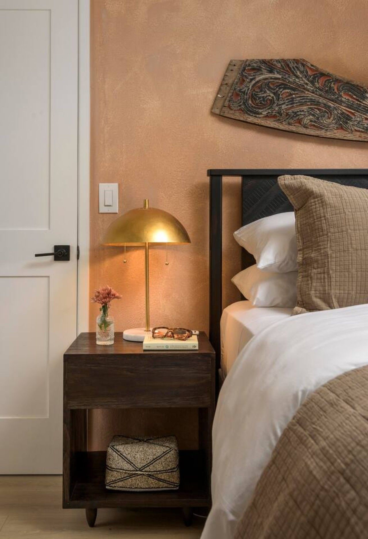 A neatly made bed with white bedding, a bedside lamp, a small bedside table with a vase and book, and a wall-mounted artwork.