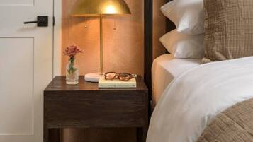 A neatly made bed with white bedding, a bedside lamp, a small bedside table with a vase and book, and a wall-mounted artwork.