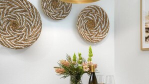 A dining area with a round wooden table, wicker chairs, a hanging light fixture, and woven wall decorations.