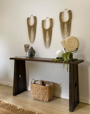 A wooden console table with a woven basket underneath, a Buddha statue, and three wall-mounted woven wall hangings. - Private Balcony | Coastal Escape (Summerland)