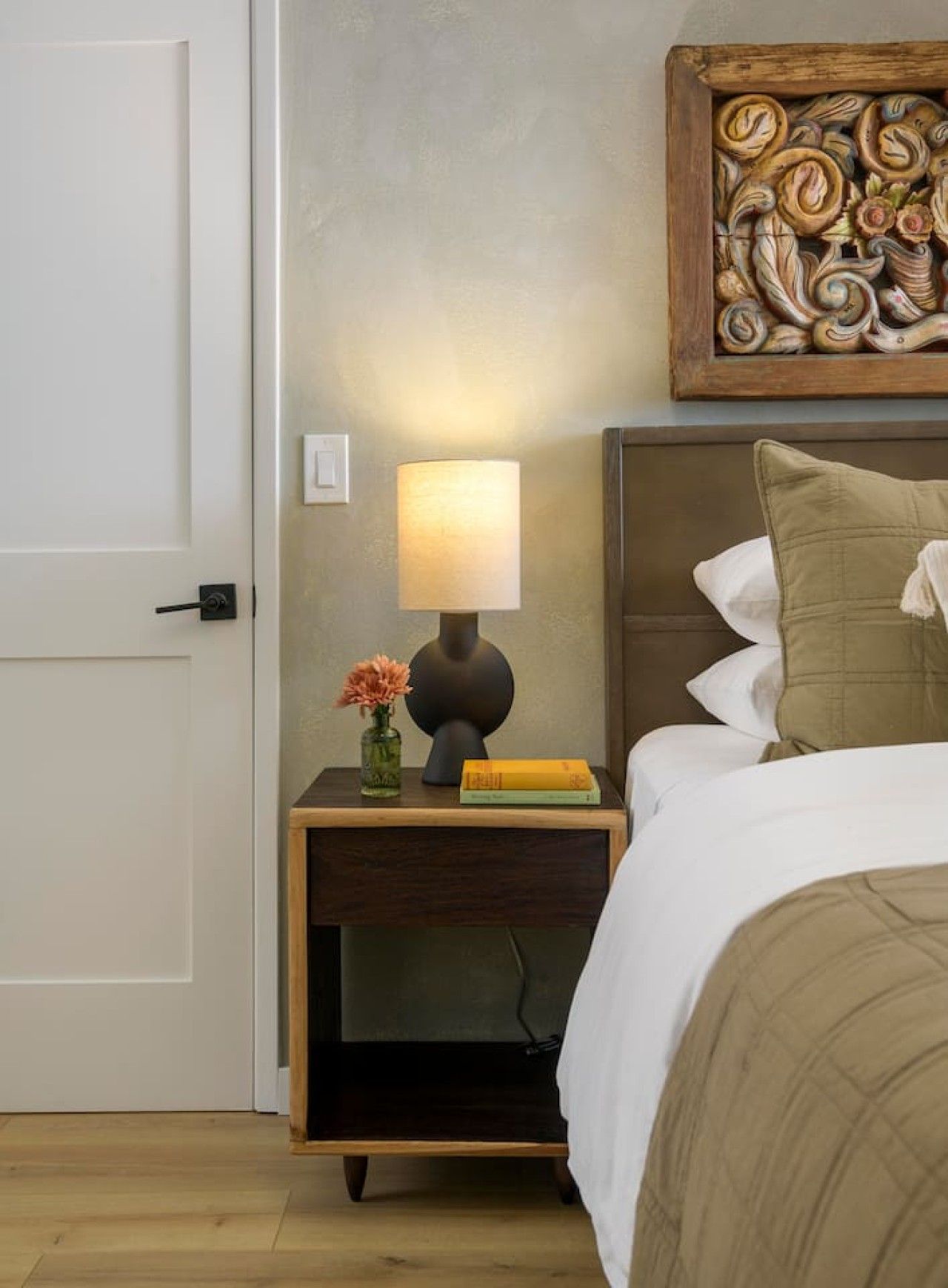 A bedroom with a bed, a bedside table with a lamp, a decorative piece above the bed, and a door.