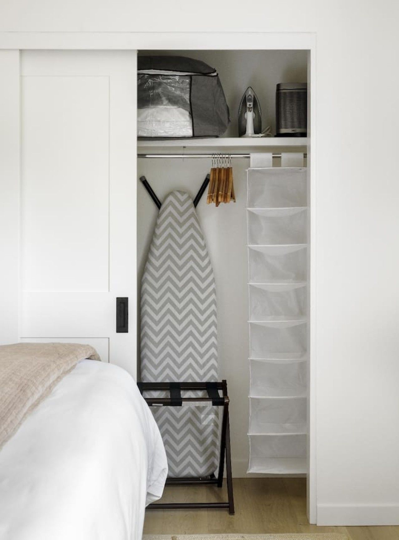 Een slaapkamer met een bed, een strijkplank, een kledingrek en een inloopkast met planken.