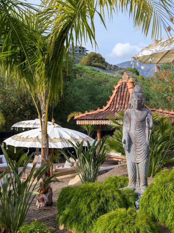 Un jardín con una estatua de Buda, palmeras y una estructura de techo tradicional.