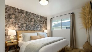 A bedroom with a bed, nightstands, a window with curtains, and a wall with a tropical wallpaper. - Group-Friendly | Tranquil Oasis (Summerland)
