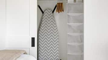 A bedroom with a bed, an ironing board, a hanging rack, and a closet with shelves.
