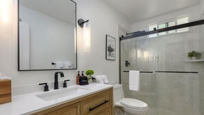 A modern bathroom with a large mirror, a white sink, a black faucet, wooden cabinets, a toilet, and a glass shower enclosure. - Group-Friendly | Tranquil Oasis (Summerland)