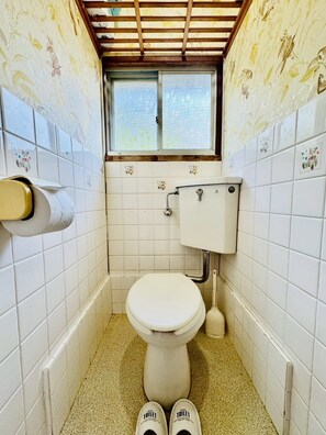 Une petite salle de bain étroite, équipée de toilettes, d’une fenêtre et d’un porte-rouleau de papier toilette.