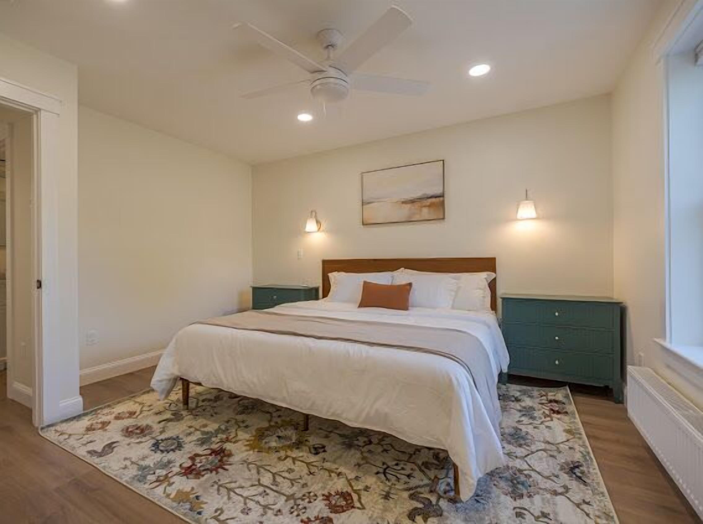 A bedroom with a bed, a ceiling fan, a nightstand, a lamp, and a window.