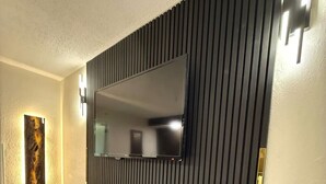 Un salon moderne avec un téléviseur à écran plat fixé sur un mur foncé orné de lambris verticaux, un meuble de rangement noir et une petite table ronde surmontée d’une lampe.