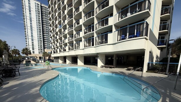 Une piscine dont la profondeur est indiquée comme étant de 3 pieds et 6 pouces, entourée d’un bâtiment doté de nombreux balcons et d’un espace extérieur aménagé pour s’asseoir.