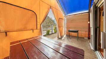 Un espacio al aire libre cubierto con una mesa de madera y un banco, vista a follaje a través de una ventana, y barandilla de metal.
