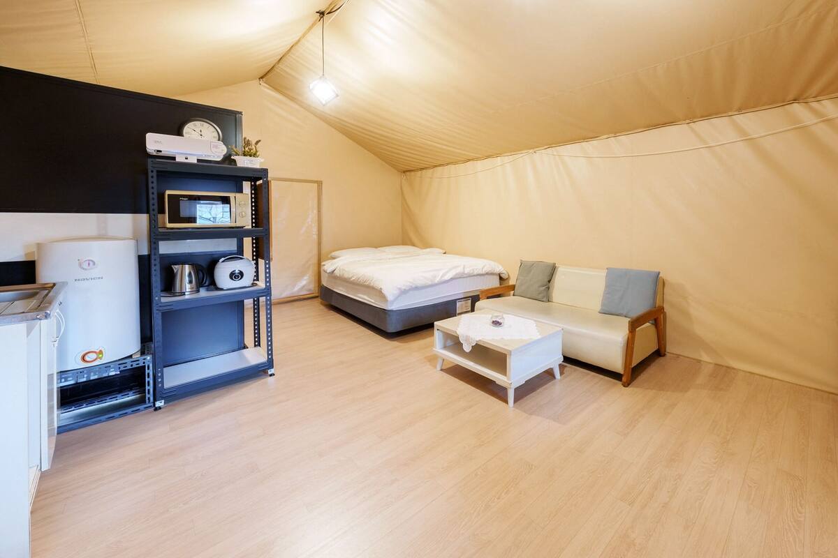Une pièce aménagée comme une tente, avec un lit, un canapé, une petite table, un t éléviseur et une étagère contenant des appareils électroménagers.