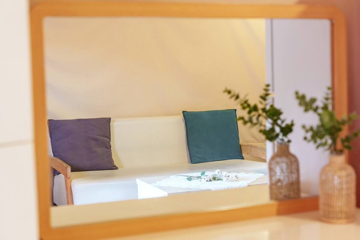Un miroir reflétant un canapé avec deux coussins, une petite table recouverte d’une nappe en dentelle et deux vases avec des plantes.