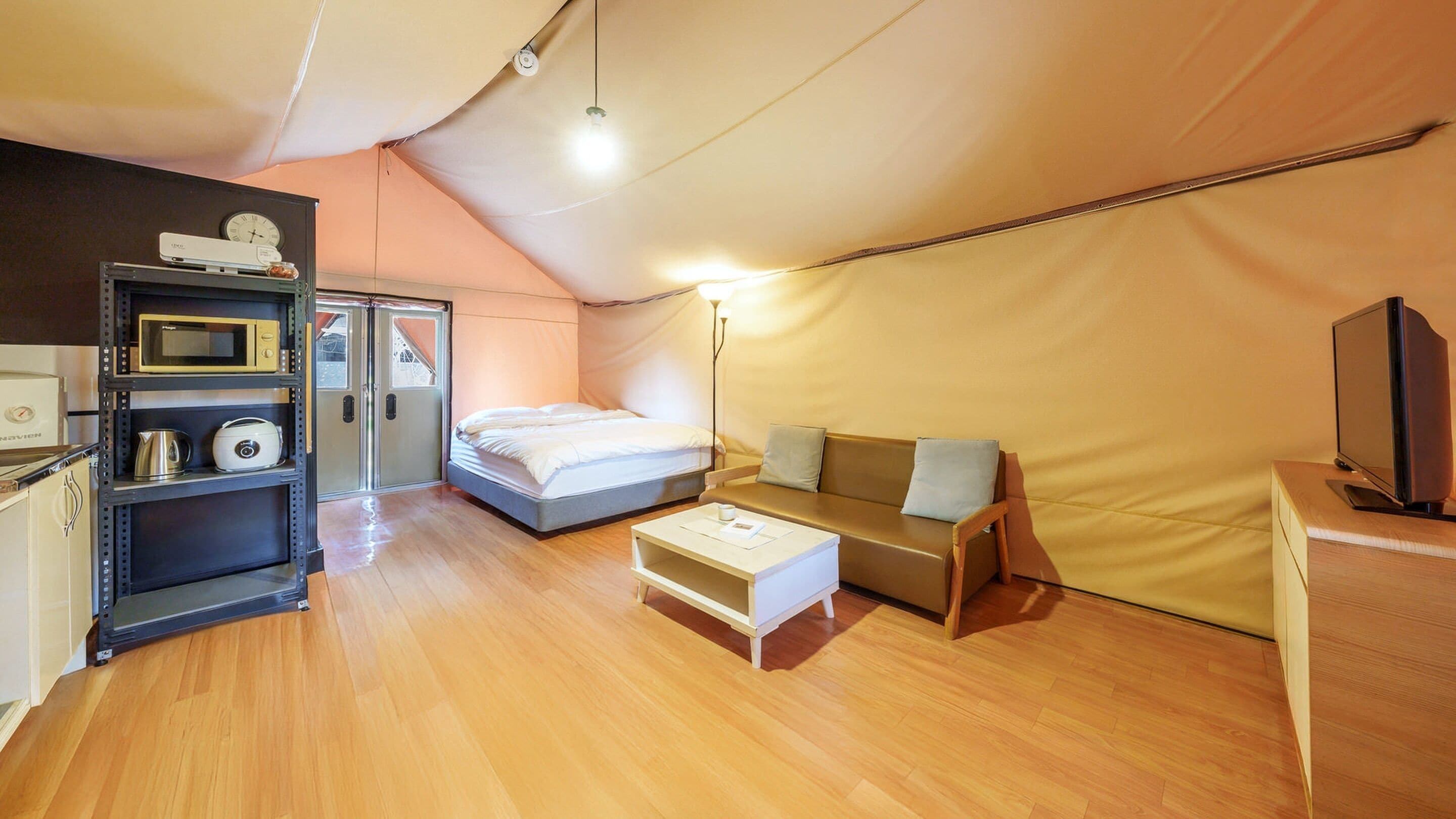 A compact room with a bed, a sofa, a coffee table, a TV, and a kitchenette with a microwave and kettle.