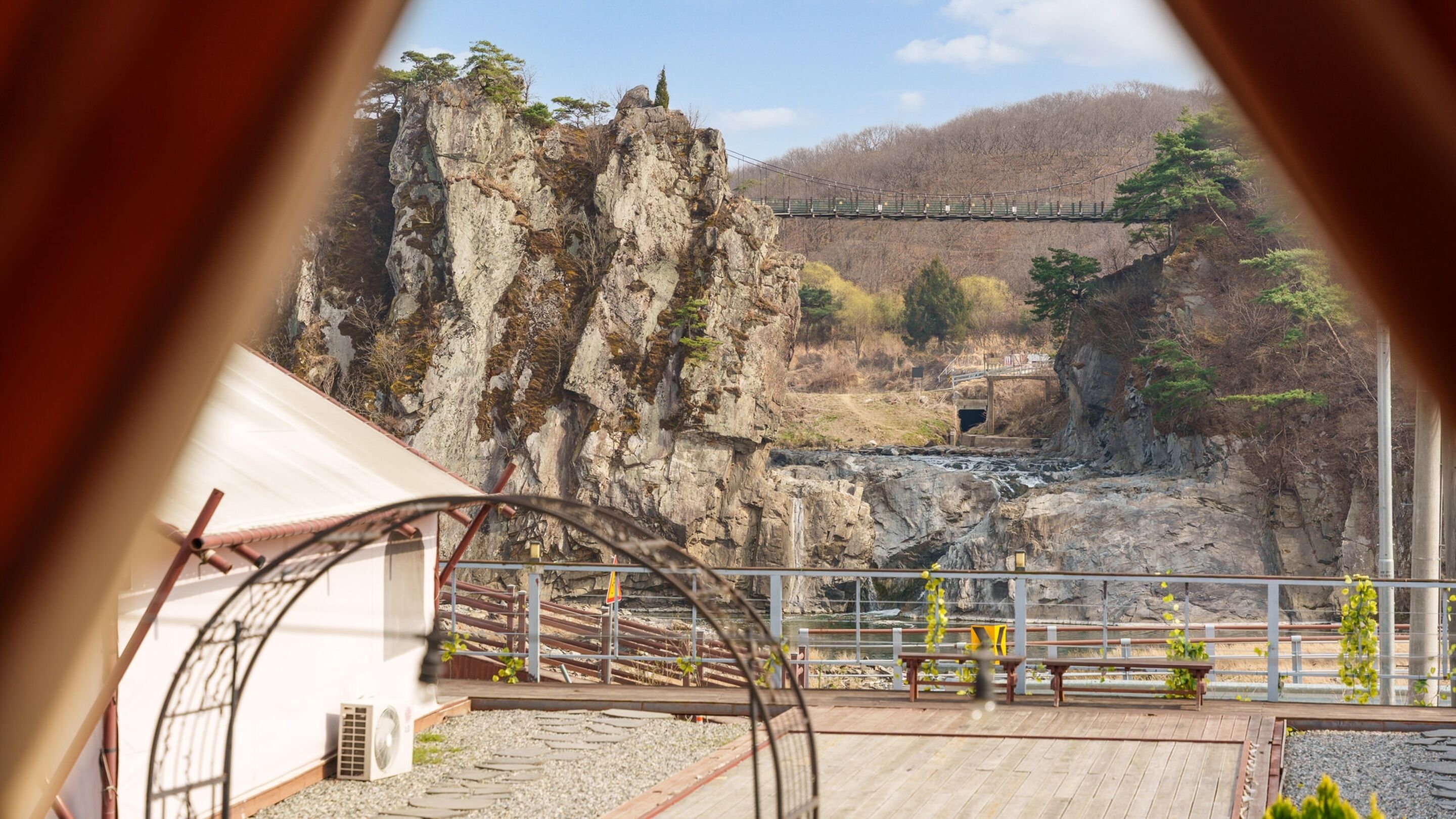 A view of a rocky cliff with a suspension bridge, a wooden deck, and a small waterfall.