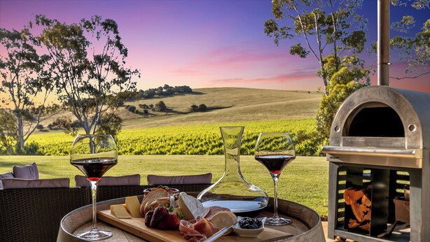 An outdoor dining setup with a wooden barrel labeled 170121, a pizza, and two glasses of red wine.