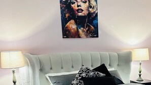 A bedroom with a bed, two nightstands, a lamp on each, a large painting on the wall, and a white throw blanket on the bed.
