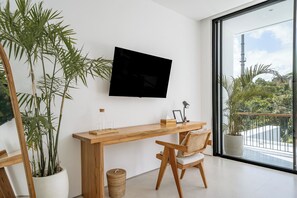 A modern room with a wooden desk, a chair, a television mounted on the wall, and large windows offering a view of greenery outside. - Elements A3 Villa Bingin (Pecatu)