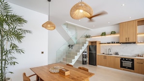 A modern kitchen with wooden cabinets, a marble countertop, and a built-in oven. There is a dining table with chairs and a potted plant. - Elements A3 Villa Bingin (Pecatu)