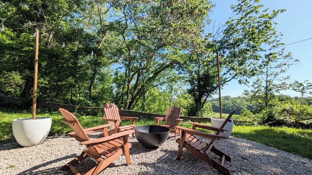 Een terras met Adirondack-stoelen en een vuurkuil, omringd door bomen en een grindpad.