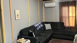 Una sala de estar con un sofá seccional oscuro, un taburete de madera, un tapete tejido y un equipo de aire acondicionado en la pared.