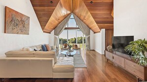 A modern living room with a wooden ceiling, a sofa, a coffee table, a TV, and a large window offering a view of the outdoors.