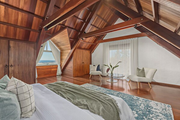 Amplia habitación en el ático con techo alto, vigas de madera, una cama grande, una zona de estar con una silla y una mesa, y vistas a través de una ventana.
